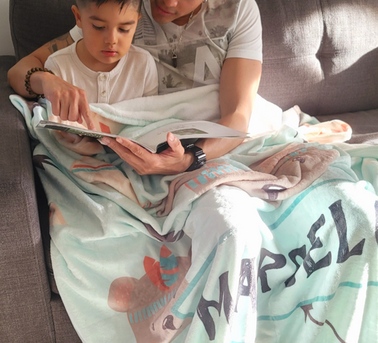 father reading a book to his son sitting on the sofa in the living room while covering their laps with a personalized baby fleece blanket from Hunny Bubba kids