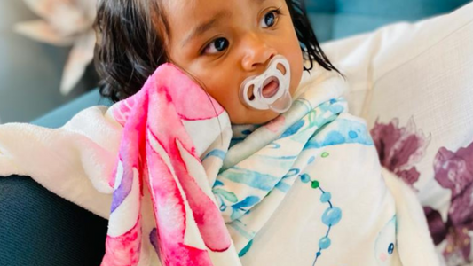 baby girl sitting on a sofa sucking on a pacifier while she is covered with a soft and warm personalized baby fleece blanket from Hunny Bubba Kids