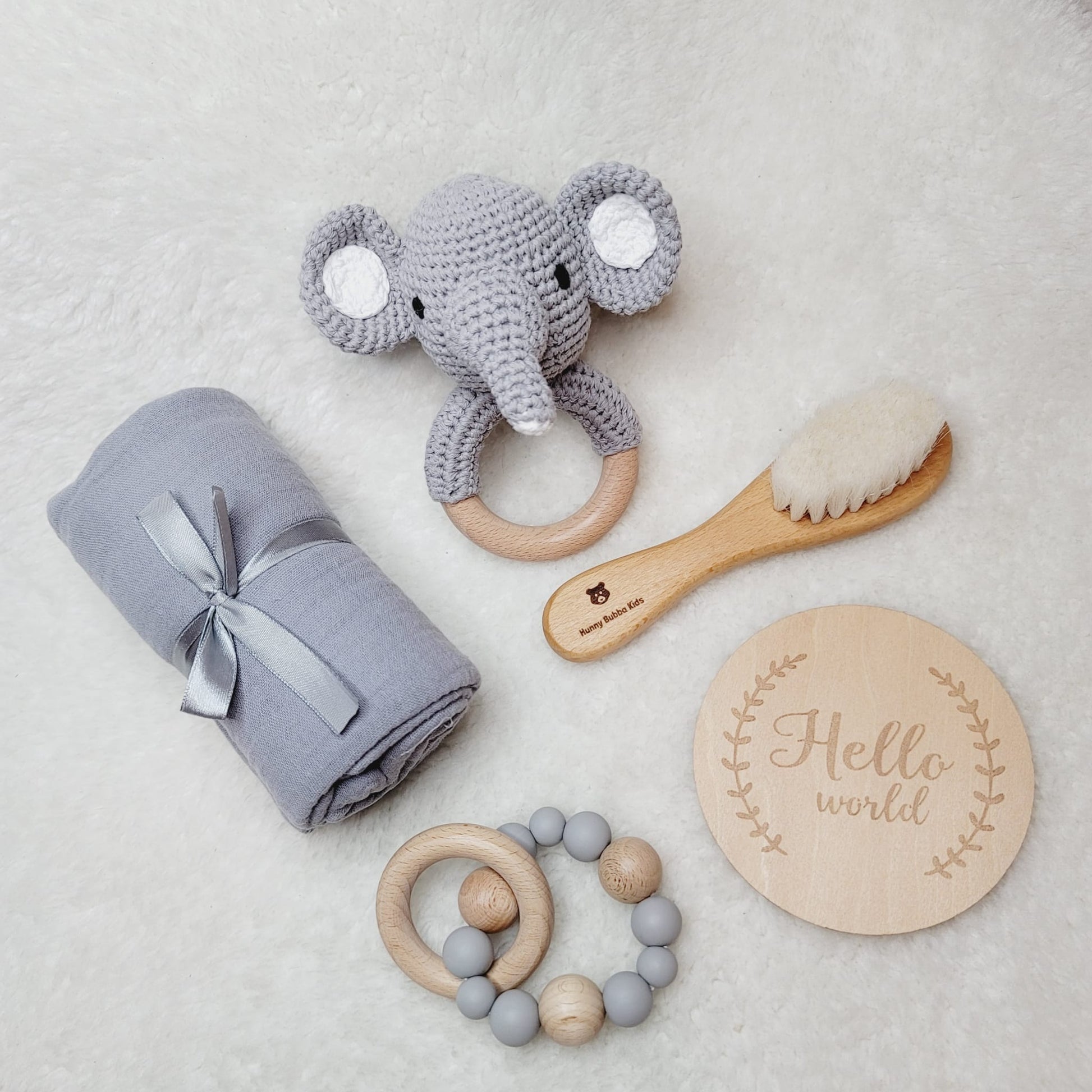 a grey newborn baby gift set with an elephant crochet rattle, a hairbrush, and wooden and silicone rattle and a swaddle laid on a soft surface
