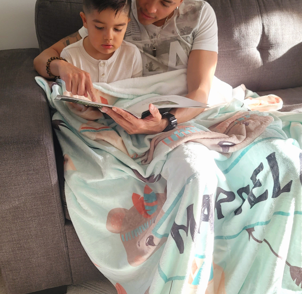 father and son spending some quality time reading a book and using a warm personalized baby fleece blanket to cover themselves.