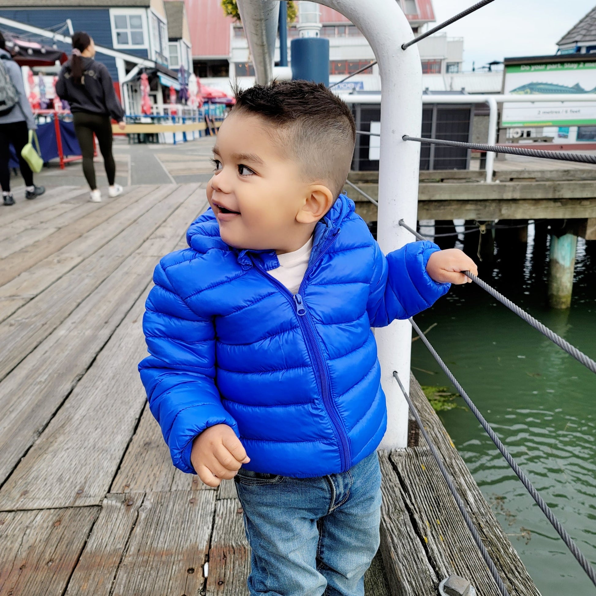Baby boy wearing a blue kids puffer jacket on a deck by the sea