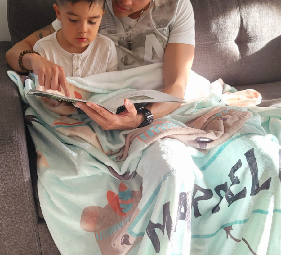 father reading a book to his son sitting on the sofa in the living room while covering their laps with a personalized baby fleece blanket from Hunny Bubba kids