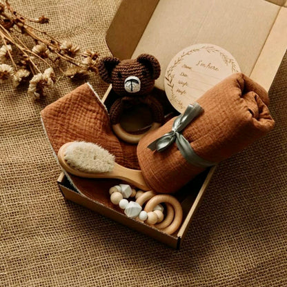a brown newborn baby gift set with a brown bear crochet rattle, a hair brush, a swaddle, a bib and a wooden and silicone teething toy packed in a box.