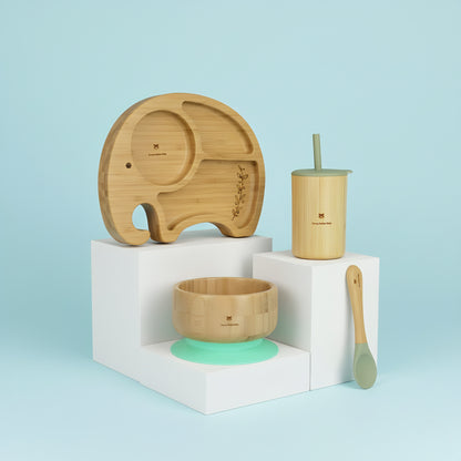 Set of bamboo baby utensils including a plate, cup, bowl, and spoon on a light blue background.
