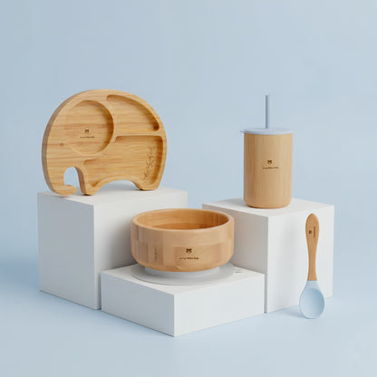 Set of wooden dining utensils for babies including a plate, bowl, cup, and spoon on a light blue background.