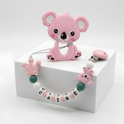 Pink koala-shaped teether attached to personalized pacifier clip with a chain and baby's name on it on a white background