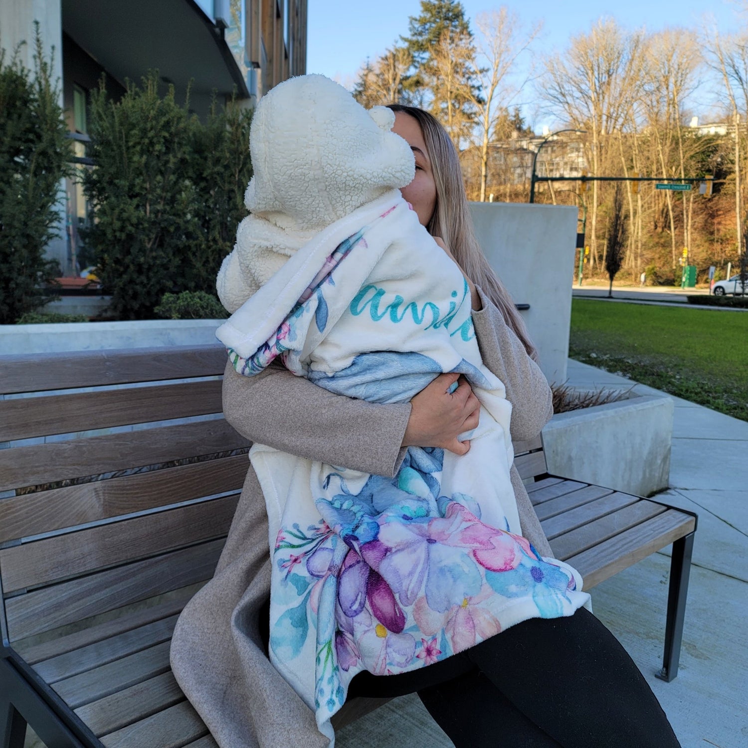 Mom sitting on a bench outside holding her baby in her arms while covering the baby with a personalized fleece baby blanket with name on it 