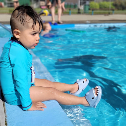 toddler boy sitting down on edge of pool wearing blue shark slippers 