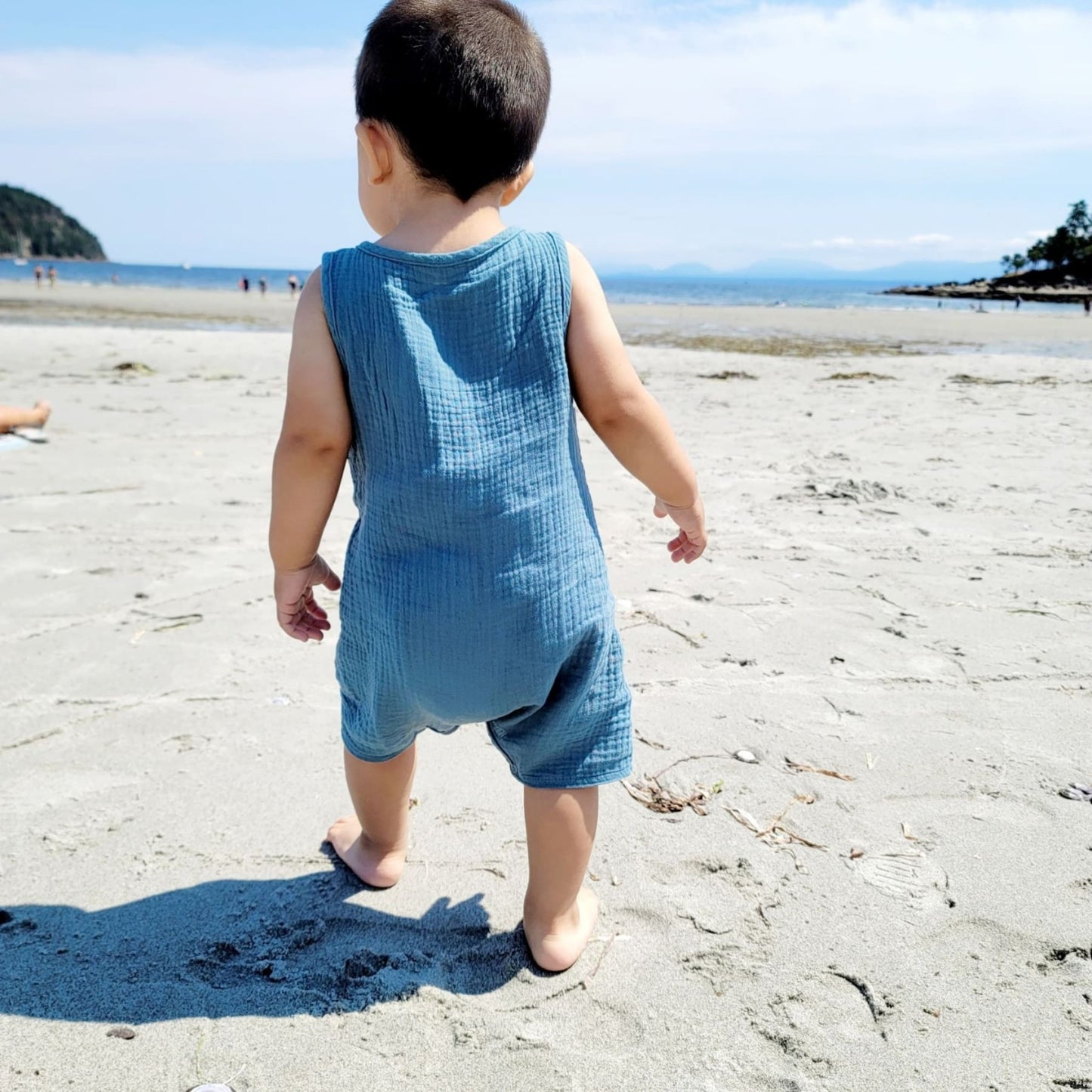 The back side of a baby wearing a cute blue romper at the beach- hunny bubba kids